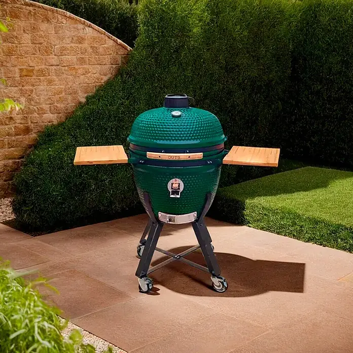 Outr Ahumador Kamado Grill XL - Diámetro de la superficie de la parrilla: 51,5 cm, Verde - Parrilla kamado de cerámica verde con estantes laterales de madera sobre soporte con ruedas, en un patio rodeado de setos verdes.