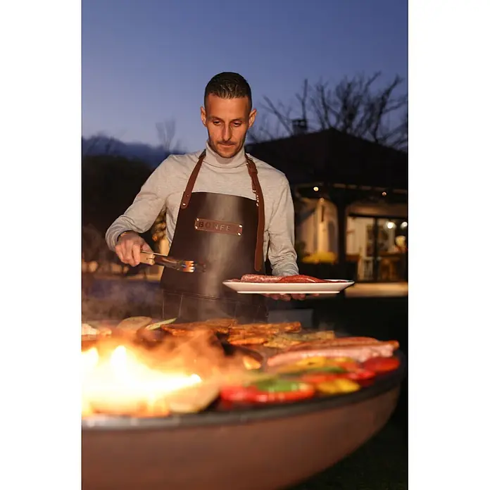 Bonfeu Brasero barbacoa Bonbiza - Ø x Al: 98 x 102 cm, Acero, Marrón óxido - Hombre con delantal Bonfeu de cuero marrón oscuro asando al aire libre, con pinzas y plato de salchichas.