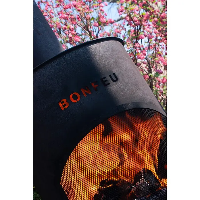 Bonfeu Chimenea de exterior Bonton - Ø x Al: 60 x 195 cm, Acero, Negro - Fosa de fuego de metal gris oscuro con recorte "BONFEU", mostrando llamas vibrantes a través de una malla. Fondo borroso de árbol en flor rosa.