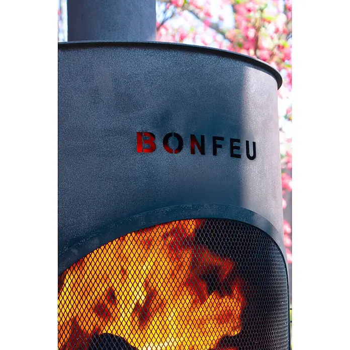 Bonfeu Chimenea de exterior Bonton - Ø x Al: 60 x 195 cm, Acero, Negro - Chimenea de exterior Bonfeu gris oscuro con acabado texturizado, mostrando llamas vibrantes a través de una malla protectora, con flores rosas borrosas.