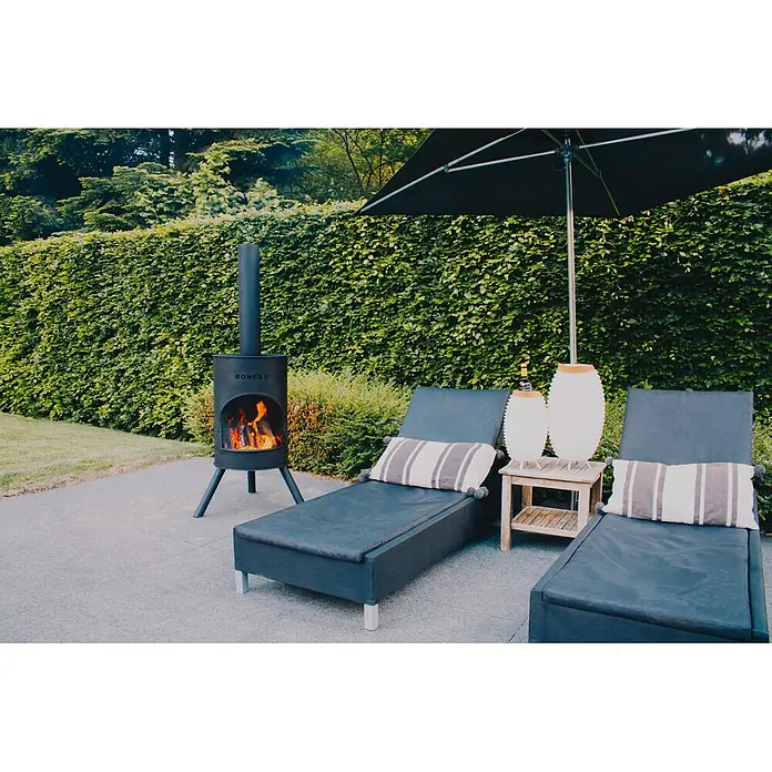 Bonfeu Chimenea de exterior Bonton - Ø x Al: 60 x 195 cm, Acero, Negro - Chimenea de exterior de metal negro con fuego encendido, junto a tumbonas bajo una sombrilla.