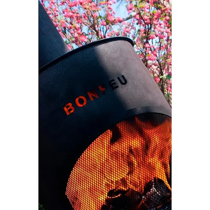 Bonfeu Chimenea de exterior Bonton - Ø x Al: 50 x 172 cm, Acero, Negro - Fogonero exterior negro BONFEU con fuego brillante visible a través de la malla y el nombre de la marca recortado, con árboles de flores rosadas de fondo.