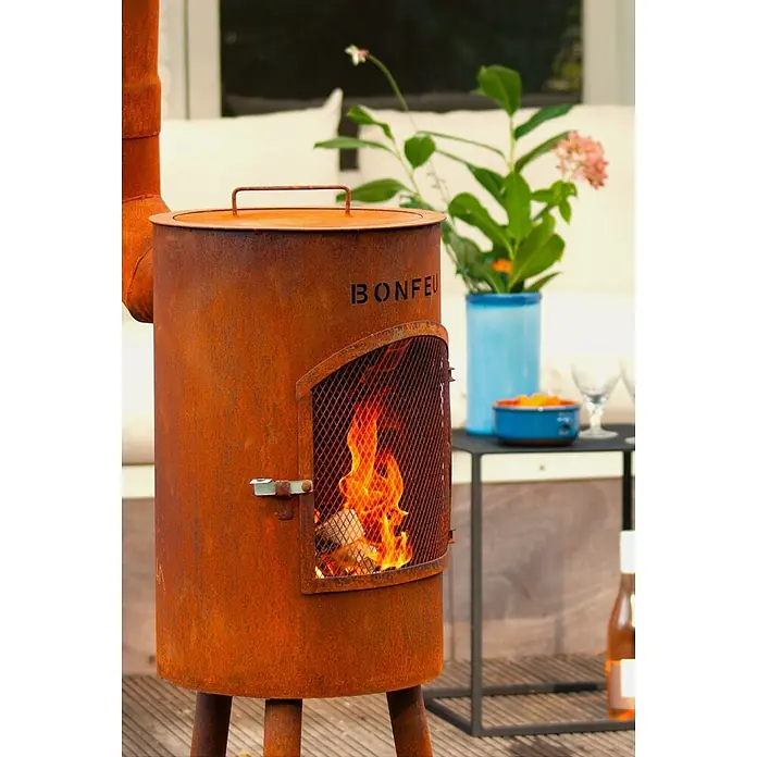 Bonfeu Chimenea de exterior Bongiano S - Ø x Al: 30 x 142 cm, Acero Corten, Marrón óxido - Estufa exterior oxidada con fuego ardiendo visible a través de la puerta de malla, situada en un patio.
