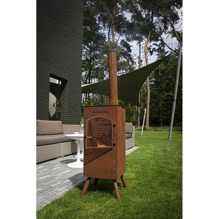 Bonfeu Chimenea de exterior Boncarre - L x An x Al: 35 x 35 x 164 cm, Acero Corten, Marrón óxido - Chimenea exterior de metal oxidado (BONFEU) con fuego encendido en un jardín.