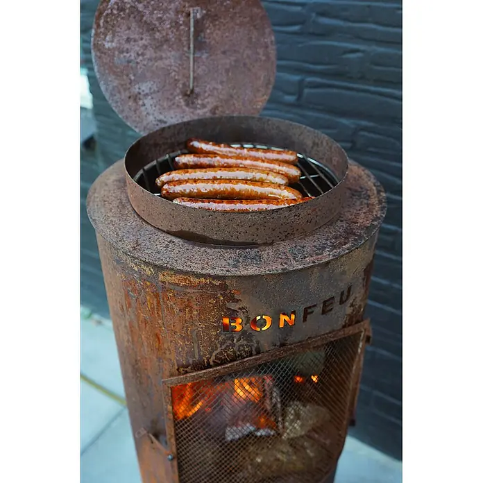 Bonfeu Chimenea de exterior Bontino L - Ø x Al: 35 x 164 cm, Acero Corten, Marrón óxido - Barbacoa BONFEU rústica de metal oxidado con salchichas asándose y fuego visible abajo.