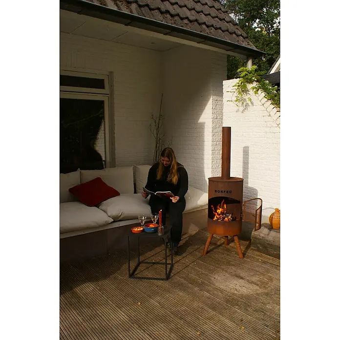 Bonfeu Chimenea de exterior Bonton - Ø x Al: 40 x 145 cm, Acero Corten, Marrón óxido - Una chimenea de exterior rústica de acero corten con fuego encendido, creando ambiente en un patio donde una persona lee.