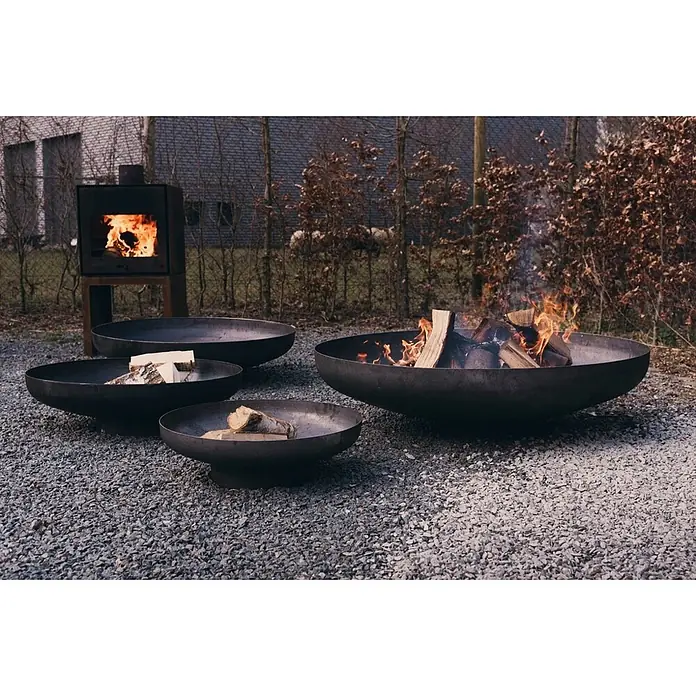 Outr Brasero Jesco - Diámetro: 55 cm, Acero Corten - Cuatro braseros de metal oscuro sobre grava. Uno arde con leña; los otros contienen leña sin encender. Una estufa exterior también arde.