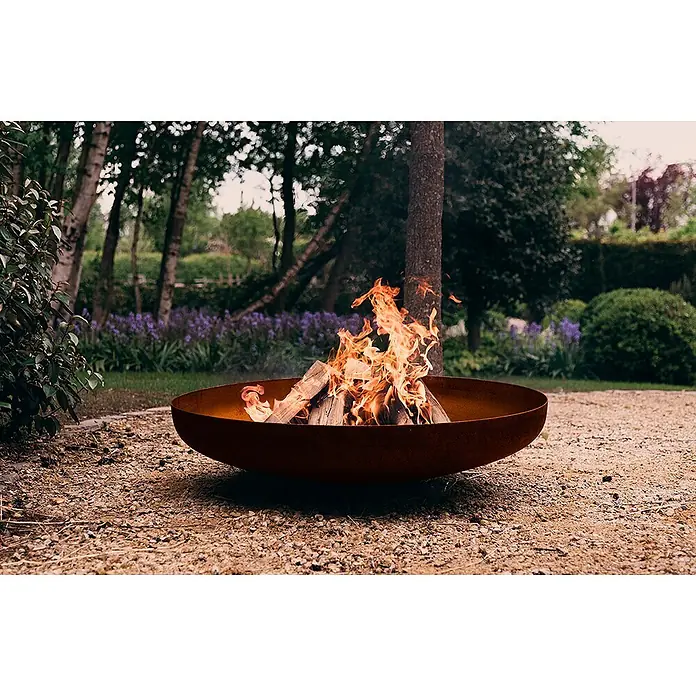 Outr Brasero Jesco - Diámetro: 55 cm, Acero Corten - Un fogón redondo de metal rústico con fuego ardiente, en un patio de grava en un jardín exuberante.