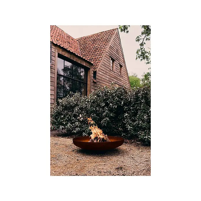 Outr Brasero Jesco - Diámetro: 55 cm, Acero Corten - Brasero de metal oxidado con llamas, en el exterior frente a una casa rústica de madera.