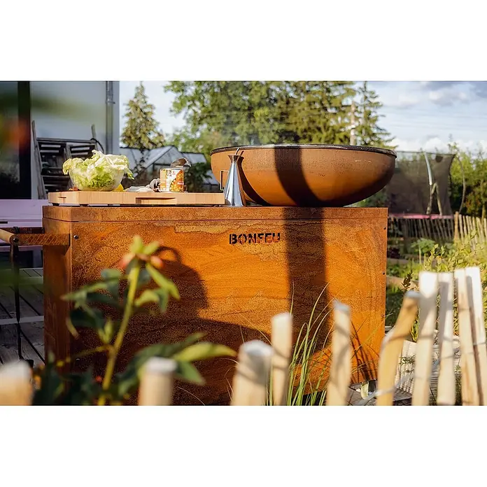 Bonfeu Brasero barbacoa Bonbiza Island - Ø x Al: 82 x 110 cm, Acero, Marrón óxido - Unidad de cocción exterior BONFEU de metal oxidado con gran parrilla en forma de cuenco, con alimentos en un jardín.