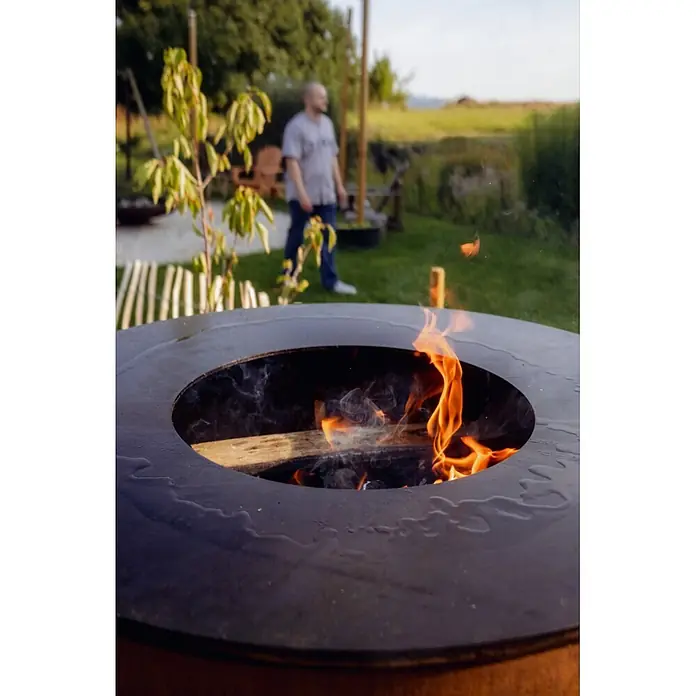 Bonfeu Brasero barbacoa Bonbiza Island - Ø x Al: 82 x 110 cm, Acero, Marrón óxido - Fogonero de metal oscuro con fuego ardiente y humo, al aire libre. Una persona está borrosa en el fondo.