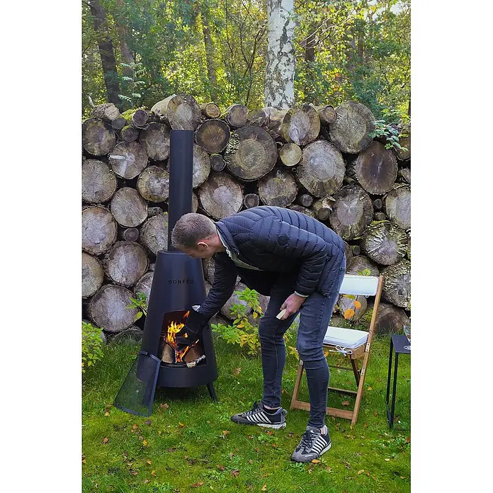 Bonfeu Chimenea de exterior Bonpyra - Ø x Al: 50 x 166 cm, Acero, Negro - Foso de fuego cónico negro Bonfeu siendo usado al aire libre con un hombre añadiendo leña al fuego.