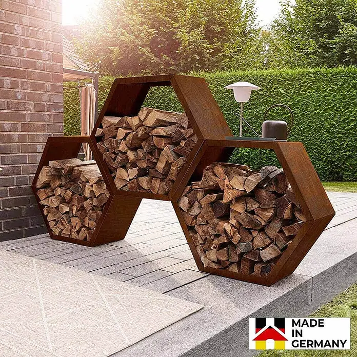 Home Deluxe Holzlege ANTARES Menge: 3 -  - Hexagonales, rostfarbenes Metall-Kaminholzregal mit Holz gefüllt, auf einer Terrasse stehend.