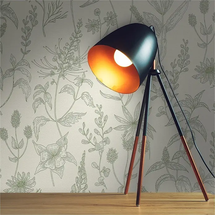 Tadessi Vliestapete M1-No.11447 - Weiß, Floral - Eine schwarz-kupferne Dreibein-Stehlampe mit sichtbarer Glühbirne, die eine botanisch gemusterte Tapete und eine Holzoberfläche warm beleuchtet.