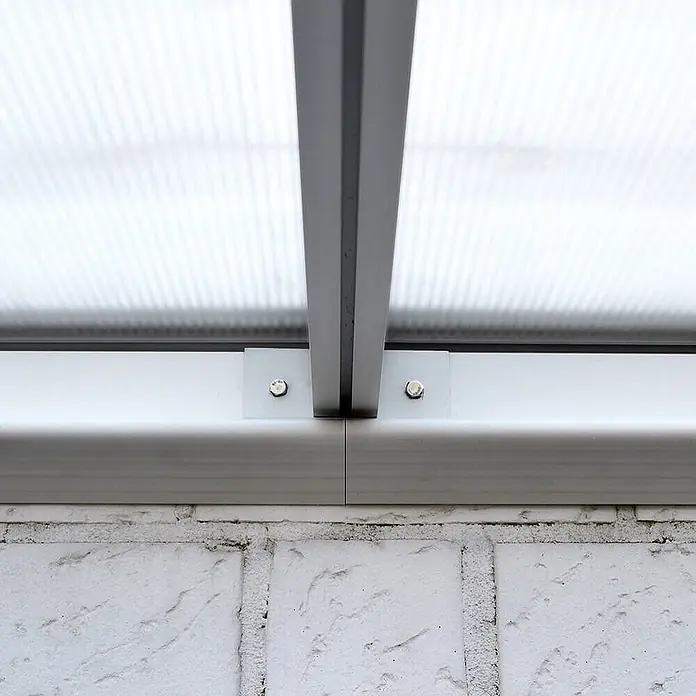 HOME DELUXE - Terrassenüberdachung SOLIS | Farbe: Weiß, Größe: 496 / 503 x 300 / 303 x 278 / 226 cm -  - Nahaufnahme einer durchsichtigen Polycarbonat-Dachplatte, mit klaren Halterungen und Schrauben an einem weißen Aluminiumrahmen auf einer weißen Ziegelwand befestigt.