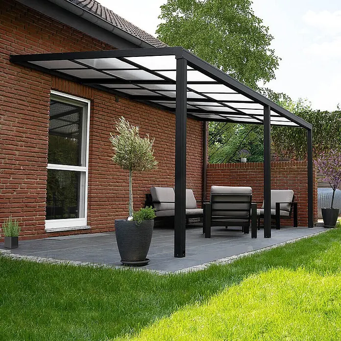 Home Deluxe Terrassenüberdachung SOLIS DELUXE Grau 390 x 295 x 233 / 272 cm -  - Schwarze Metall-Terrassenüberdachung mit lichtdurchlässigem Dach, an Ziegelhaus, über einem Sitzbereich im Freien.