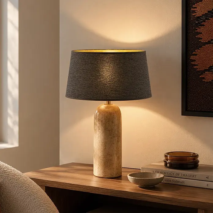 Home Sweet Home Lampenschirm Melrose - Ø x H: 35 x 19 cm, Hellgrau, Stoff, Rund - Eine Tischlampe mit hellbraunem Natursteinsockel und dunklem Stoffschirm mit goldener Innenseite, die ein warmes Licht auf einen Holztisch wirft.