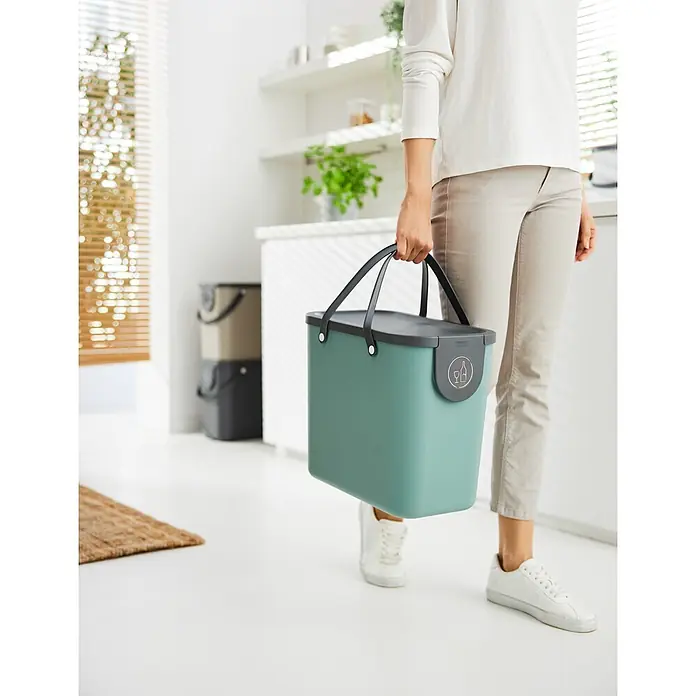 Rotho Albula Sistema de separación de residuos Albula - 25 l, Plástico, Verde muérdago - Una persona lleva un cubo de reciclaje turquesa con tapa y asas gris oscuro en una cocina moderna, ilustrando su uso para la separación de residuos.