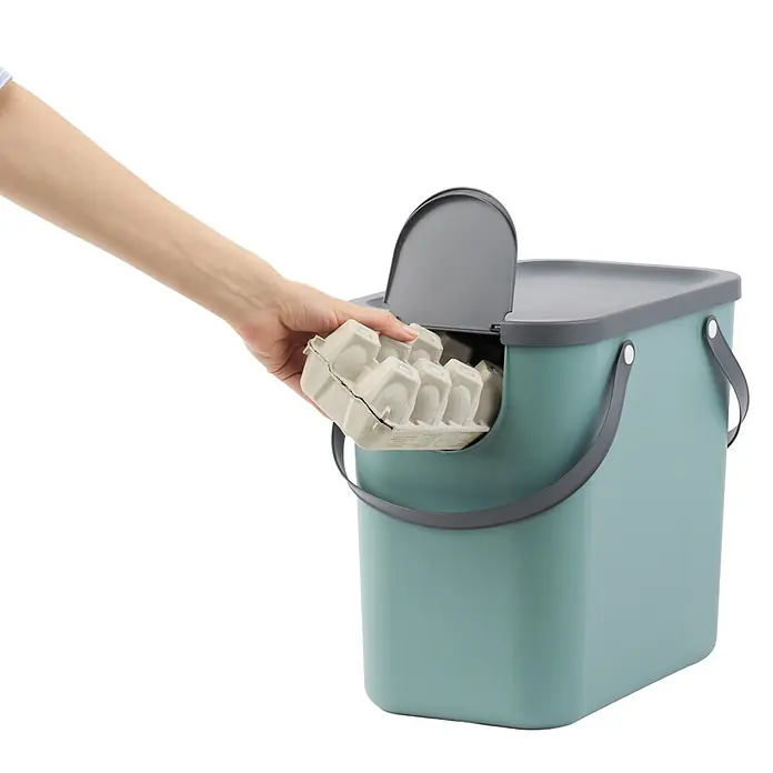 Rotho Albula Sistema de separación de residuos Albula - 25 l, Plástico, Verde muérdago - Mano colocando un cartón de huevos en la abertura lateral de un cubo de compostaje turquesa con tapa gris.