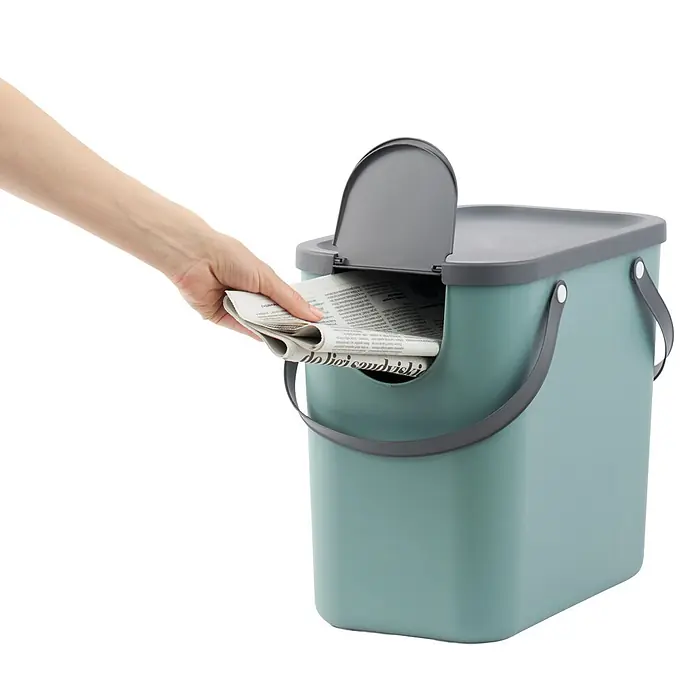 Rotho Albula Sistema de separación de residuos Albula - 25 l, Plástico, Verde muérdago - Mano colocando un periódico en un cubo de reciclaje verde-azul claro con tapa y asas grises.