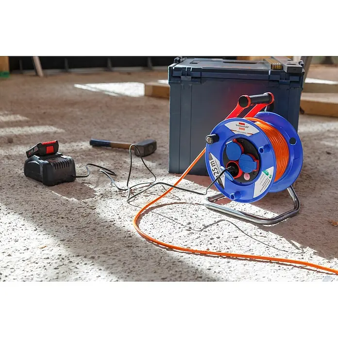 Brennenstuhl Garant Kabeltrommel - Kunststoff, 25 m, AT-N05V3V3-F, IP44 - Blaue Brennenstuhl Kabeltrommel mit orangefarbenem 25m Verlängerungskabel auf Betonboden mit Batterieladegerät und Hammer.