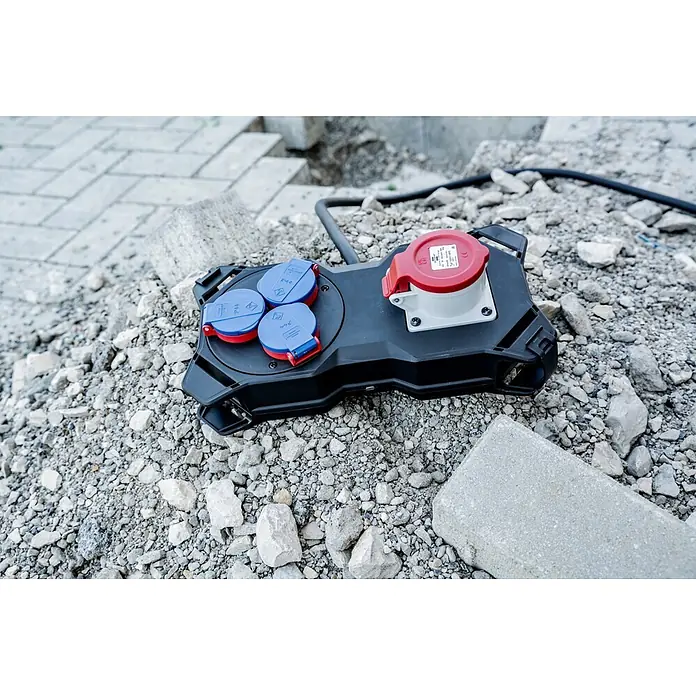 Brennenstuhl Stromverteiler Stromstation CEE IP44 - Anzahl CEE-Steckdosen: 1 Stk., Anzahl Steckdosen Schuko: 3 Stk. - Schwarzer mobiler Stromverteiler mit roter CEE-Steckdose und blauen Schuko-Steckdosen, auf Schotter einer Baustelle.