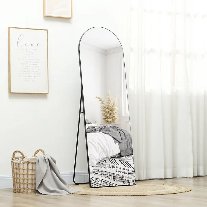 Homcom Standspiegel - Schwarz, Glas - Ein schwarz gerahmter, gewölbter Ganzkörperspiegel steht auf hellem Holzboden und reflektiert ein Bett und Pampasgras.