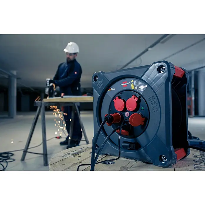 Brennenstuhl Kabeltrommel KA 3100 5 - Kunststoff, 33 m, H07RN-F, IP44 - Brennenstuhl Kabeltrommel, dunkelgrau mit roten Steckdosen, im Einsatz auf einer Baustelle mit Arbeiter im Hintergrund.
