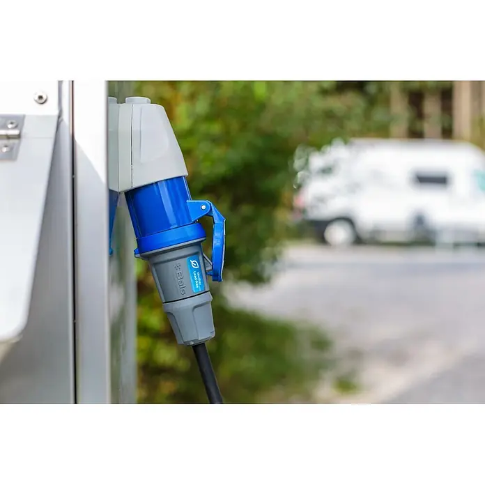Brennenstuhl Verlängerungskabel Camping-CEE - 25 m, IP44, H07RN-F - Blauer und grauer Industriestromstecker, an Außensteckdose angeschlossen, mit verschwommenem Wohnmobil im Hintergrund. Gekennzeichnet 'Bals Lead-free'.
