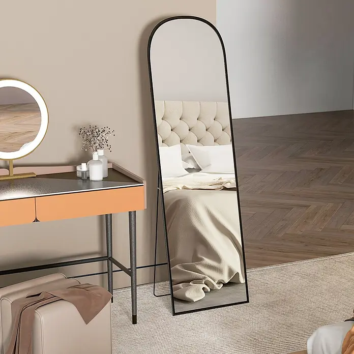 Homcom Standspiegel - Schwarz, Glas - Gebogener schwarzer Ganzkörperspiegel auf einem Teppich, der ein Bett in einem Schlafzimmer spiegelt.
