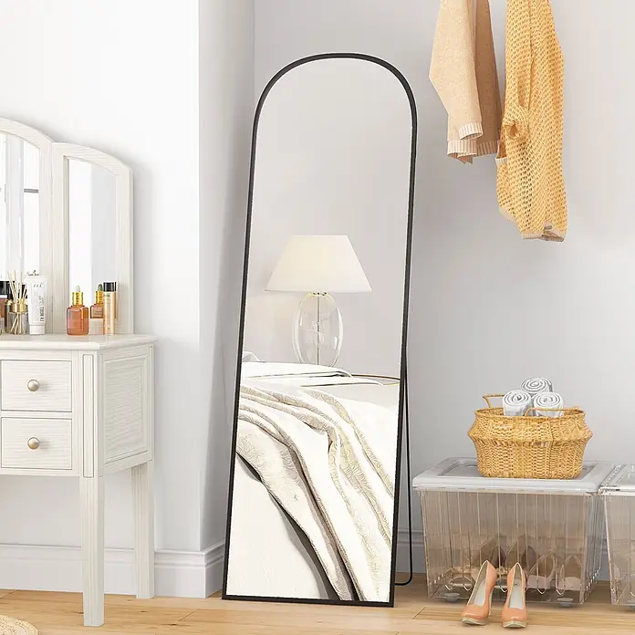 Homcom Standspiegel - Schwarz, Glas - Schwarz gerahmter, gewölbter Ganzkörperspiegel lehnt an einer Wand und spiegelt eine Schlafzimmerszene wider.