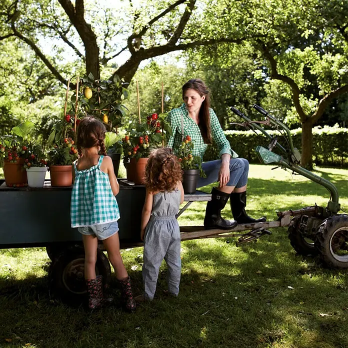 Elho Green Basics Maceta para planta - Diámetro exterior máx.: 19 cm, Plástico, Negro - Una mujer con dos niñas observa plantas de tomate en macetas en un remolque en un jardín, junto a un tractor.