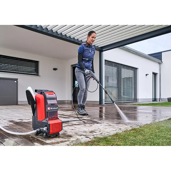 Einhell PXC 18V Akku-Hochdruckreiniger Hypresso 36/105 - 36 V, Ohne Akku - Eine Frau reinigt eine nasse Holzterrasse mit einem rot-schwarzen Einhell Hochdruckreiniger. Das kompakte Gerät steht auf dem Boden.