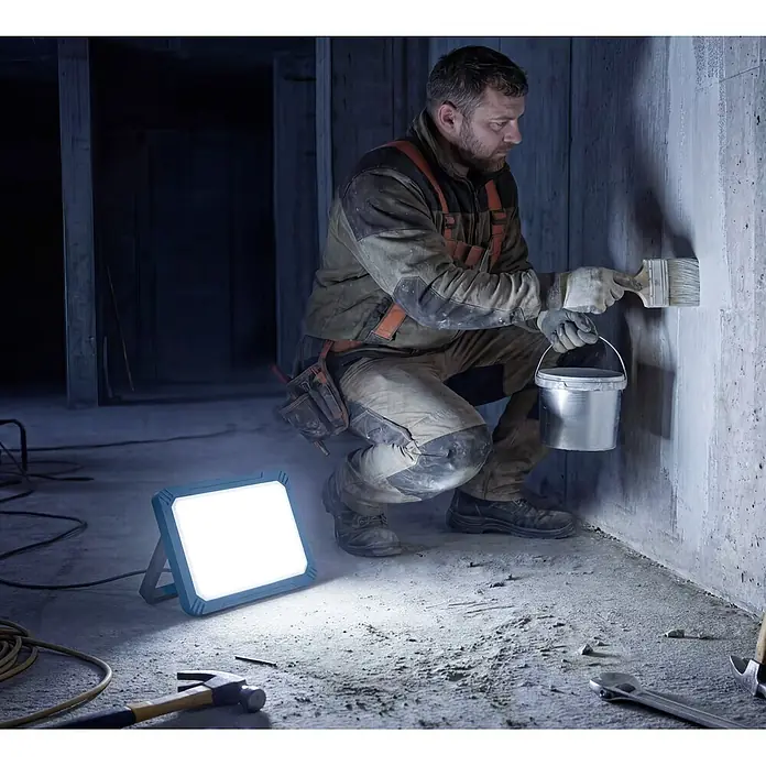 Profi Depot LED-Baustrahler - 50 W, 5.500 lm, 6.500 K, 15,5 x 21,5 x 8,7 cm, IP54 - Blauer LED-Arbeitsscheinwerfer beleuchtet einen Bauarbeiter, der eine Wand in einem dunklen Betonraum streicht.