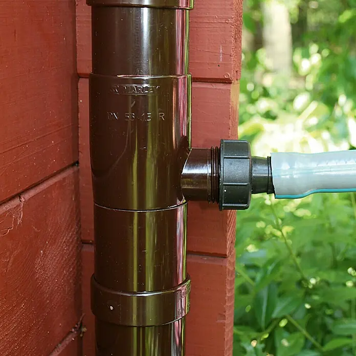 Marley Regensammler - Nennweite: 75 mm, Braun - Nahaufnahme eines braunen Regenrohrs mit seitlichem Anschluss und Verbindung zu einem grauen Schlauch an einer roten Hauswand.