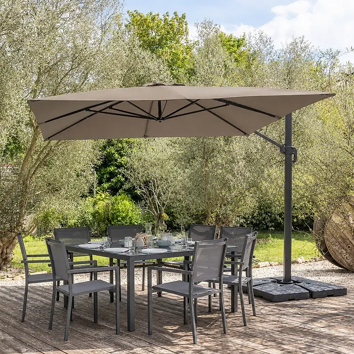 Freistehender Sonnenschirm MOLOKAI quadratisch 3x3m taupe + Hülle -  - Graues Metall-Gartenset mit Glastisch und Stühlen unter einem beigen Freiarmschirm auf einer Holzterrasse.