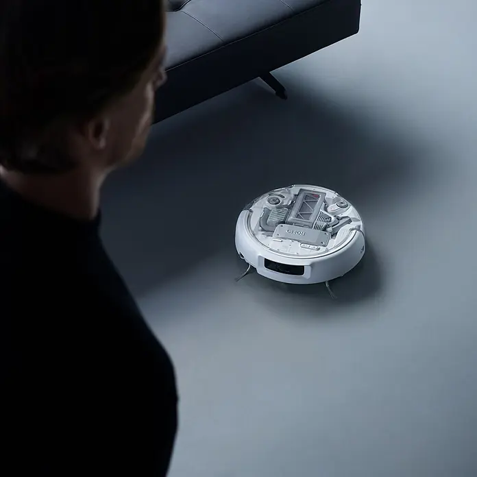 DJI Saugwischroboter P - Saugkraft: 25.000 Pa, Akkulaufzeit: 180 min, Fassungsvermögen Roboter: 260 ml - Ein weißer Saugroboter mit transparenter Oberseite, der interne Komponenten zeigt. Eine Person beobachtet ihn.