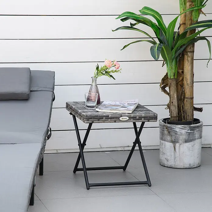 Outsunny Garten-Beistelltisch mit Stahlrahmen - Stahl, PE-Ratten, Klappbar, Quadratisch, Grau - Grauer klappbarer Beistelltisch im Rattan-Stil mit schwarzem Metallgestell, auf einer Terrasse neben einem Liegestuhl.