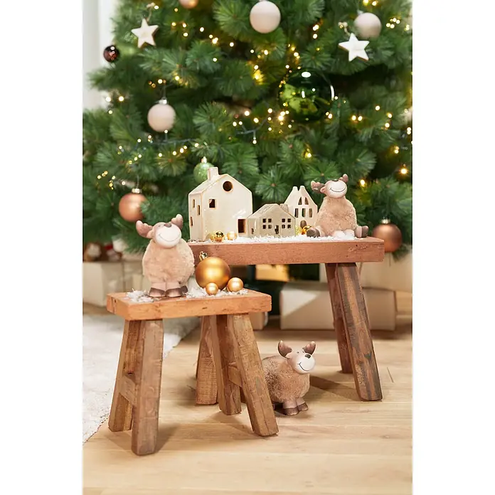 Dekoartikel LED Haus - 1 Stk., Sortiert, 10 x 8 x 14,5 cm - Zwei rustikale Holzschemel präsentieren festliche Keramikhäuser, Plüsch-Rentiere und goldene Ornamente vor einem verschwommenen Weihnachtsbaum.