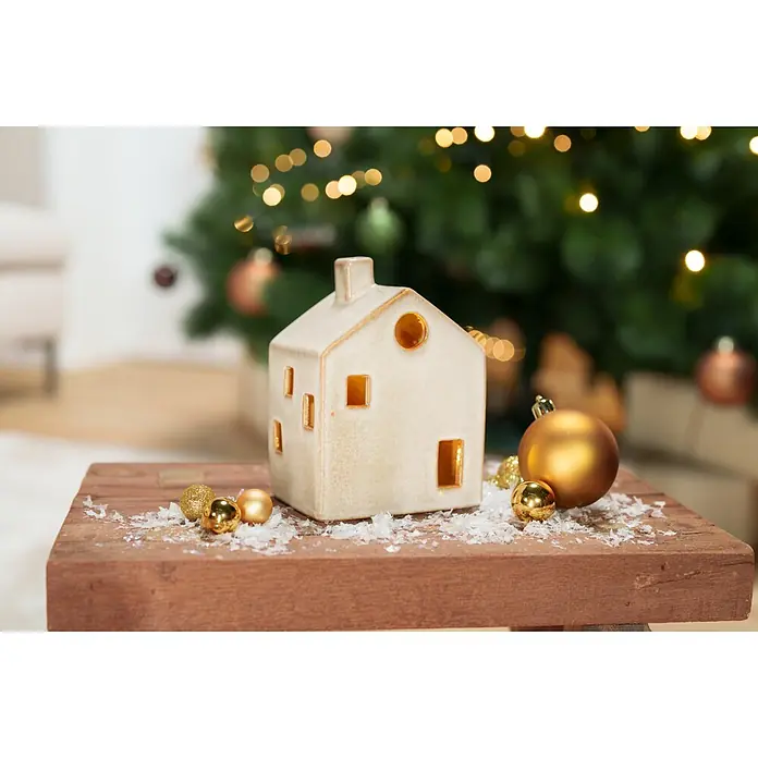 Dekoartikel LED Haus - 1 Stk., Sortiert, 10 x 8 x 14,5 cm - Cremefarbene Keramikhaus-Dekoration mit goldenen Fenstern und Ornamenten auf Holz, Weihnachtsbaum im Hintergrund.