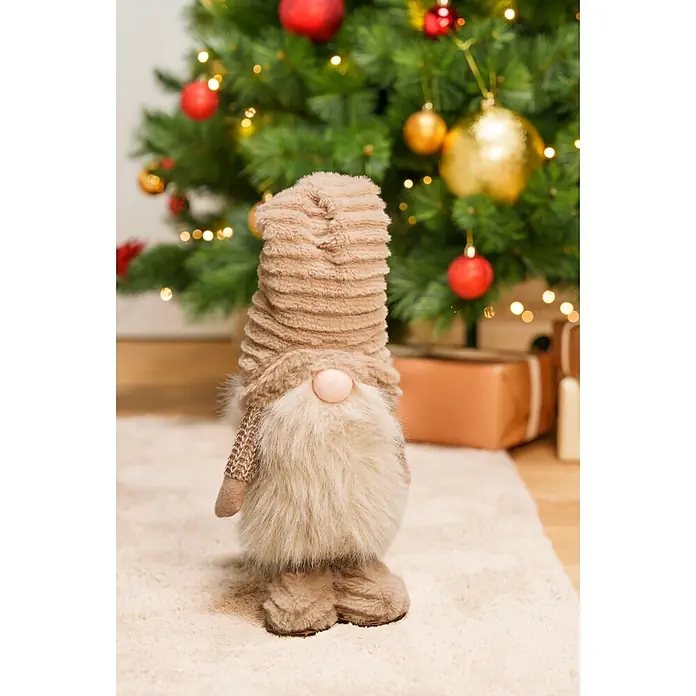 Dekofigur Weihnachtsmann - L x B x H: 18 x 13 x 65 cm, Textil - Ein braun-beiger Weihnachtswichtel mit flauschigem Bart und gerippter Mütze steht auf einem hellen Teppich vor einem geschmückten Weihnachtsbaum und Geschenken.