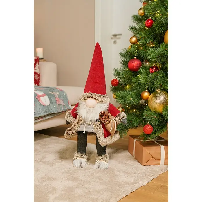 Dekofigur wackelnder Wichtel mit Jutebeutel - L x B x H: 24 x 19 x 78 cm, Textil - Festliche Weihnachtswichtel-Dekoration mit roter Mütze, weißem Bart, Pelzmantel, Kiefernzapfen und kleiner Tasche, neben geschmücktem Weihnachtsbaum.