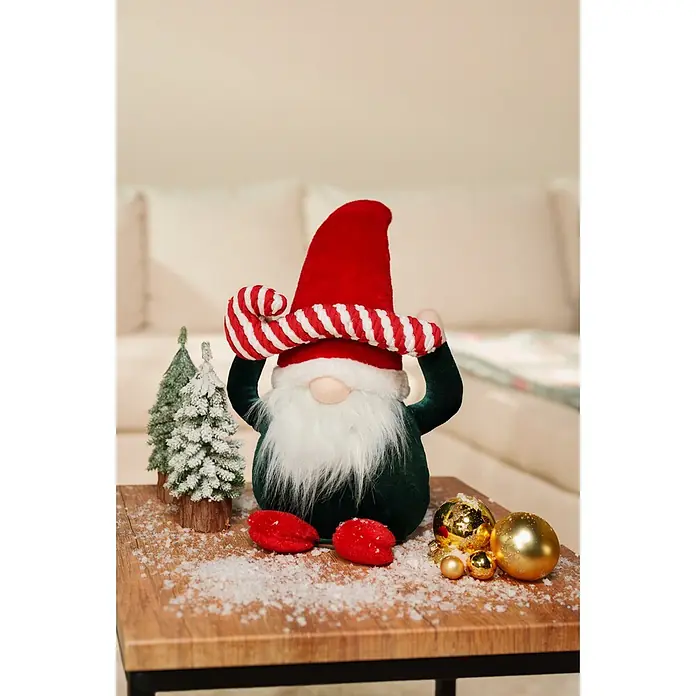 Dekofigur Weihnachtswichtel mit Zuckerstange - L x B x H: 22 x 14 x 41 cm, Polyester - Ein festlicher Weihnachtszwerg mit roter Mütze, weißem Bart, grünem Körper und roten Füßen, umgeben von verschneiten Bäumen und goldenen Kugeln auf einem Holztisch.