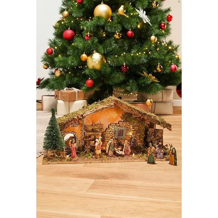 LED-Krippe Natur - 25 x 58 x 23 cm - Detaillierte Weihnachtskrippe mit Stall und Figuren, auf Holzfußboden vor einem geschmückten Weihnachtsbaum mit Geschenken.