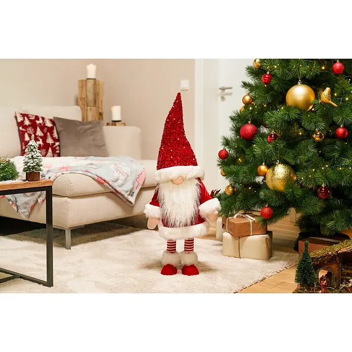 Dekofigur Weihnachtswichtel schaukelnd - L x B x H: 33 x 33 x 83 cm, Polyester - Großer Weihnachtsgnom mit roter Paillettenmütze, weißem Bart und rotem Anzug, steht neben einem geschmückten Weihnachtsbaum in einem Wohnzimmer.