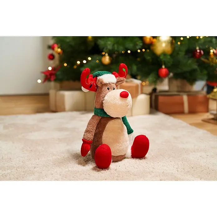 Dekofigur Reh - L x B x H: 20 x 16 x 36 cm, Polyester - Plüsch-Rentier mit roter Nase, grünem Schal und festlichem Geweih, sitzt auf einem Teppich vor einem Weihnachtsbaum.