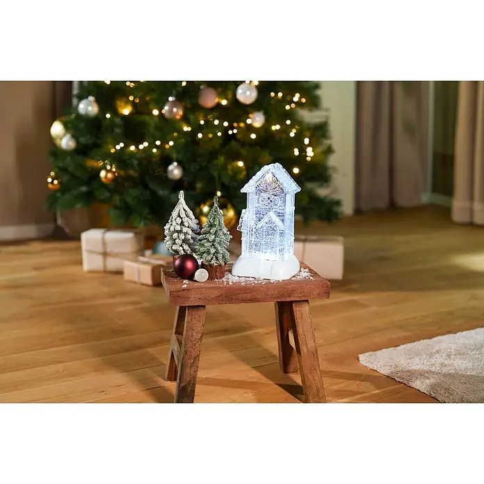 Markslöjd LED-Weihnachtsdekoleuchte Dale - 14,5 x 13 x 26 cm, Weiß/ Transparent, Batteriebetrieben, LED - Weihnachtsdekoration, darunter ein beleuchtetes Haus und beschneite Bäume, auf einem Holzschemel in einem festlichen Raum.