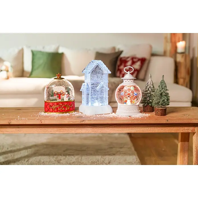 Schneekugel mit Spieluhr - Kunststoff, Weihnachtsmann - Eine Auswahl festlicher Weihnachtsdekorationen, darunter Schneekugeln, ein beleuchtetes Haus und kleine Raureifbäume, auf einem Holztisch präsentiert.