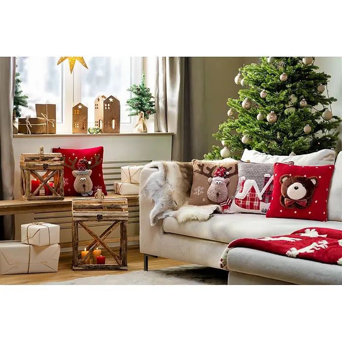 Dekokissen 3D - 40 x 40 cm, Rot/Braun - Ein festliches Wohnzimmer mit Sofa, dekoriert mit Weihnachtskissen (Rentier, Bär, Winterhaus), einem geschmückten Baum und Holzlaternen.