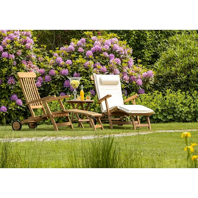 Kai Wiechmann Garten-Liegestuhl Teakholz Deckchair Europa - Teak, Klappbar, Naturbelassen - Zwei Gartenstühle aus Holz, einer mit weißen Polstern und Fußstütze, und ein kleiner Tisch mit Getränken in einem blühenden Rhododendrongarten.